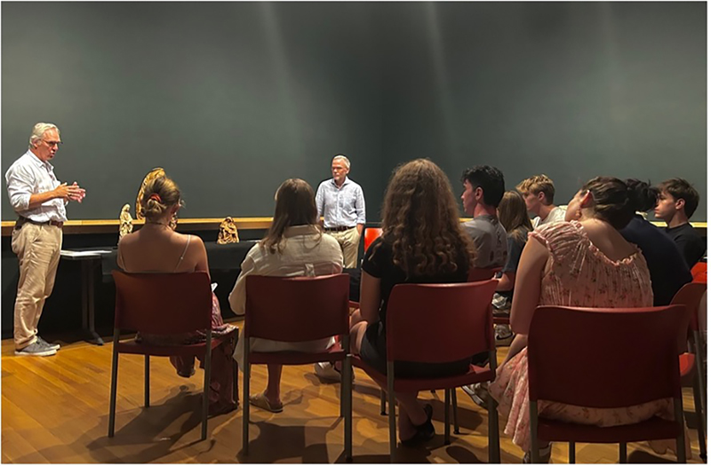 A classroom with students in chairs and two gentlemen standing and speaking to the class