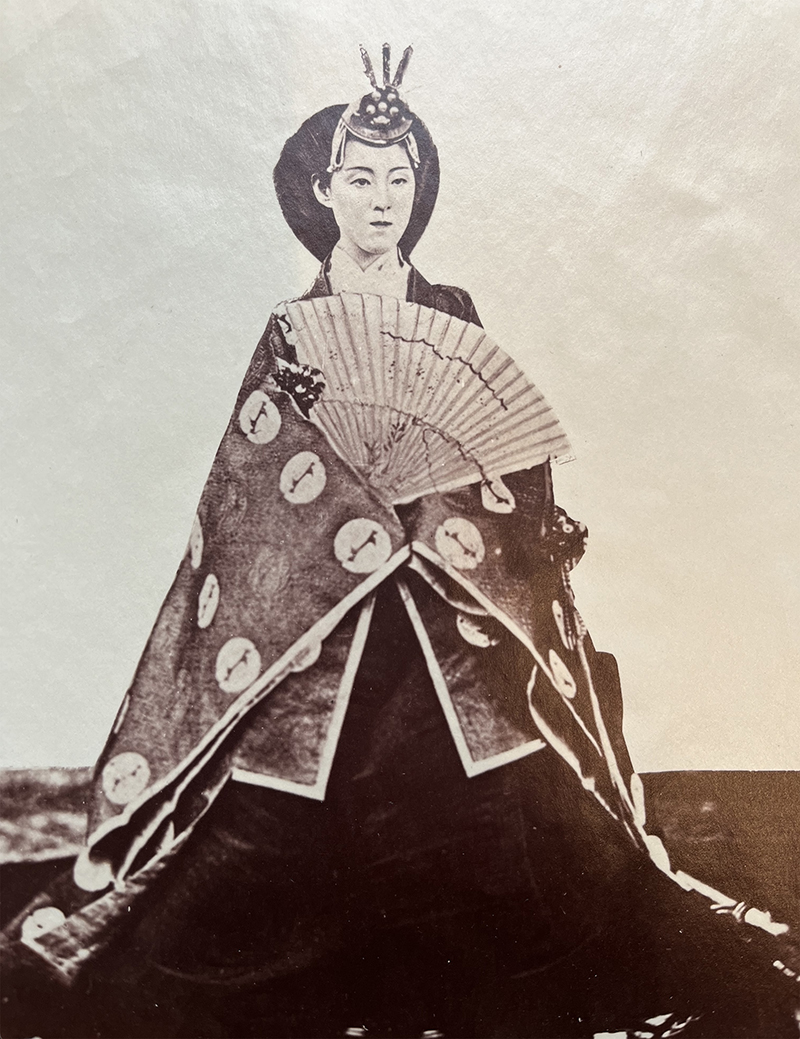 A sepia photograph of a woman in a long robe, with a fan, wearing a headpiece