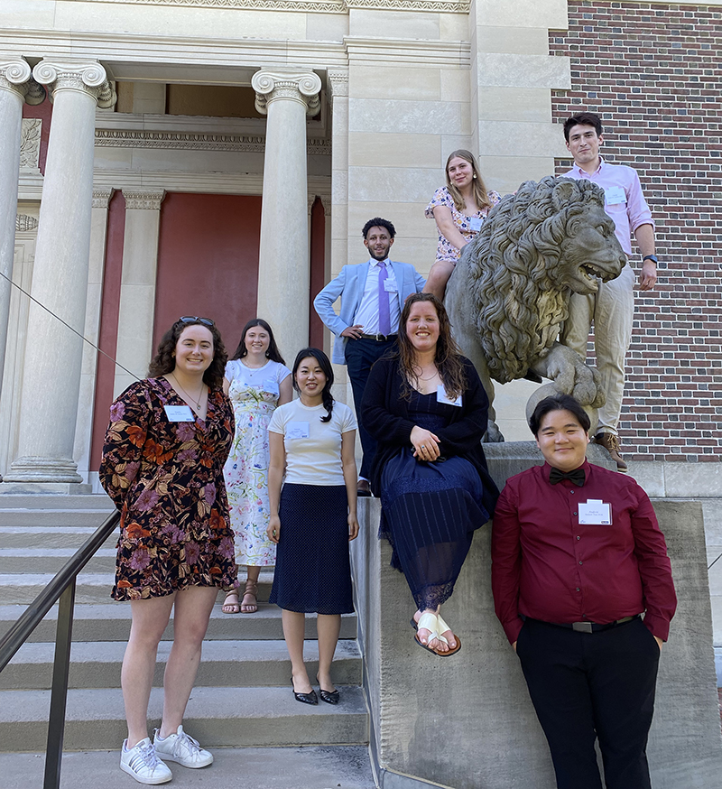 A group of 9 people posed around a statue of a lion against a brick building