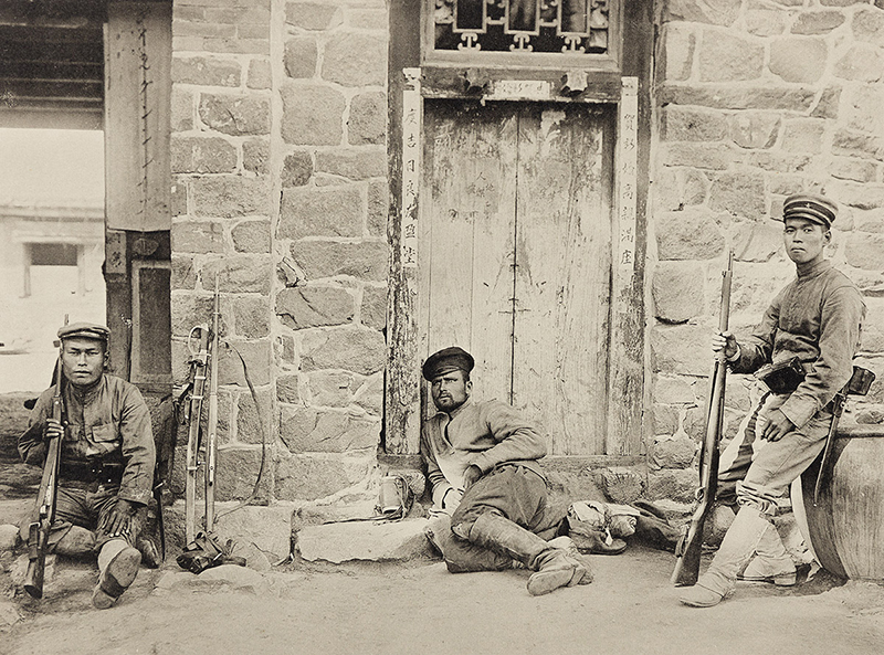 a photogravure showing three men in uniform, holding guns, in a street or courtyard