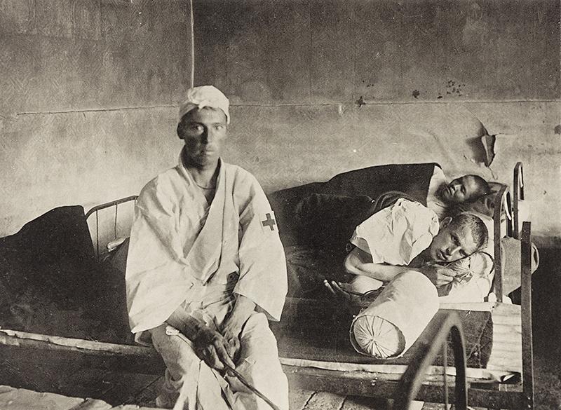 A photogravure showing two people in a bed, possibly in a hospital, and a man with a red cross on the sleeve of his garment