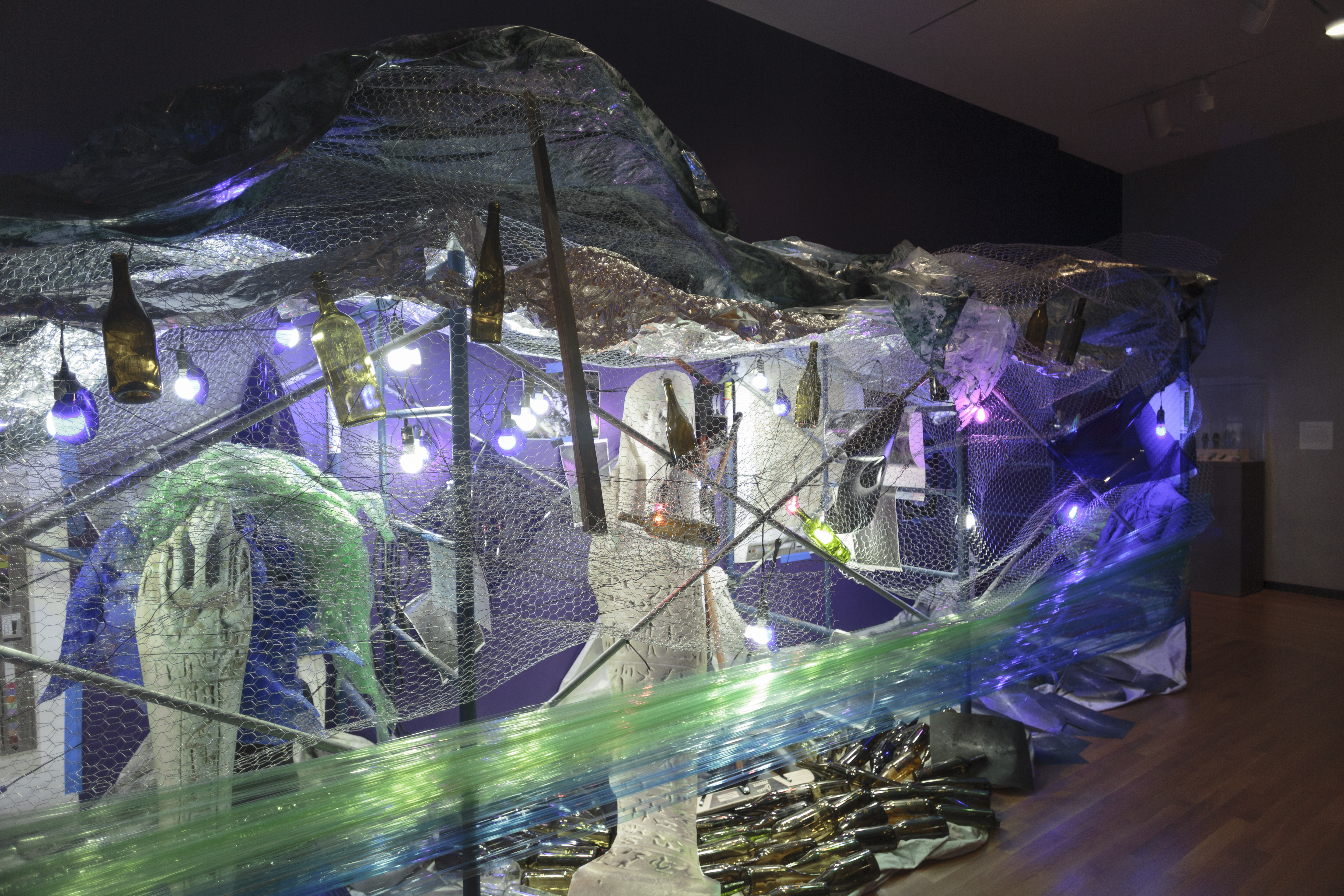 An installation artwork shows an assemblage of objects and materials illuminated by purple toned clamp lights