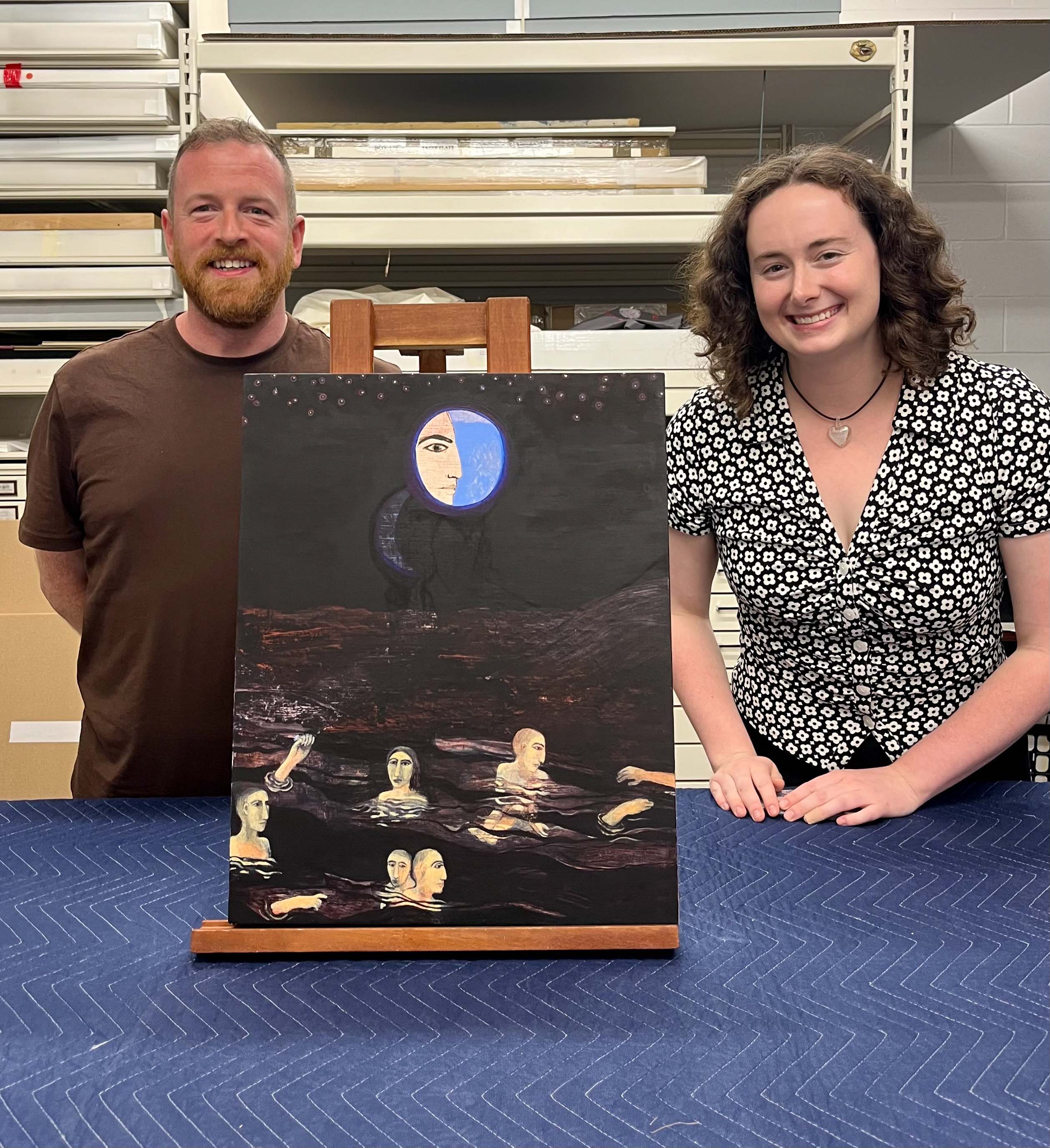 Two people stand smiling next to an unframed painting on a wooden easel