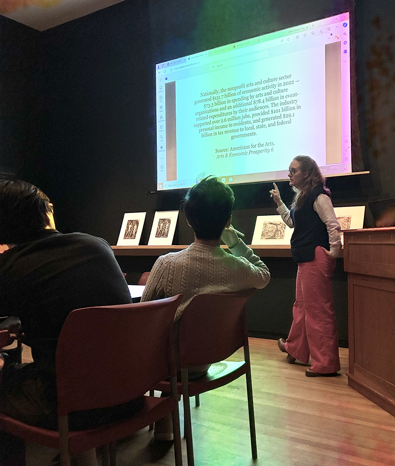 A woman in front of a screen with writing on it, speaking to students