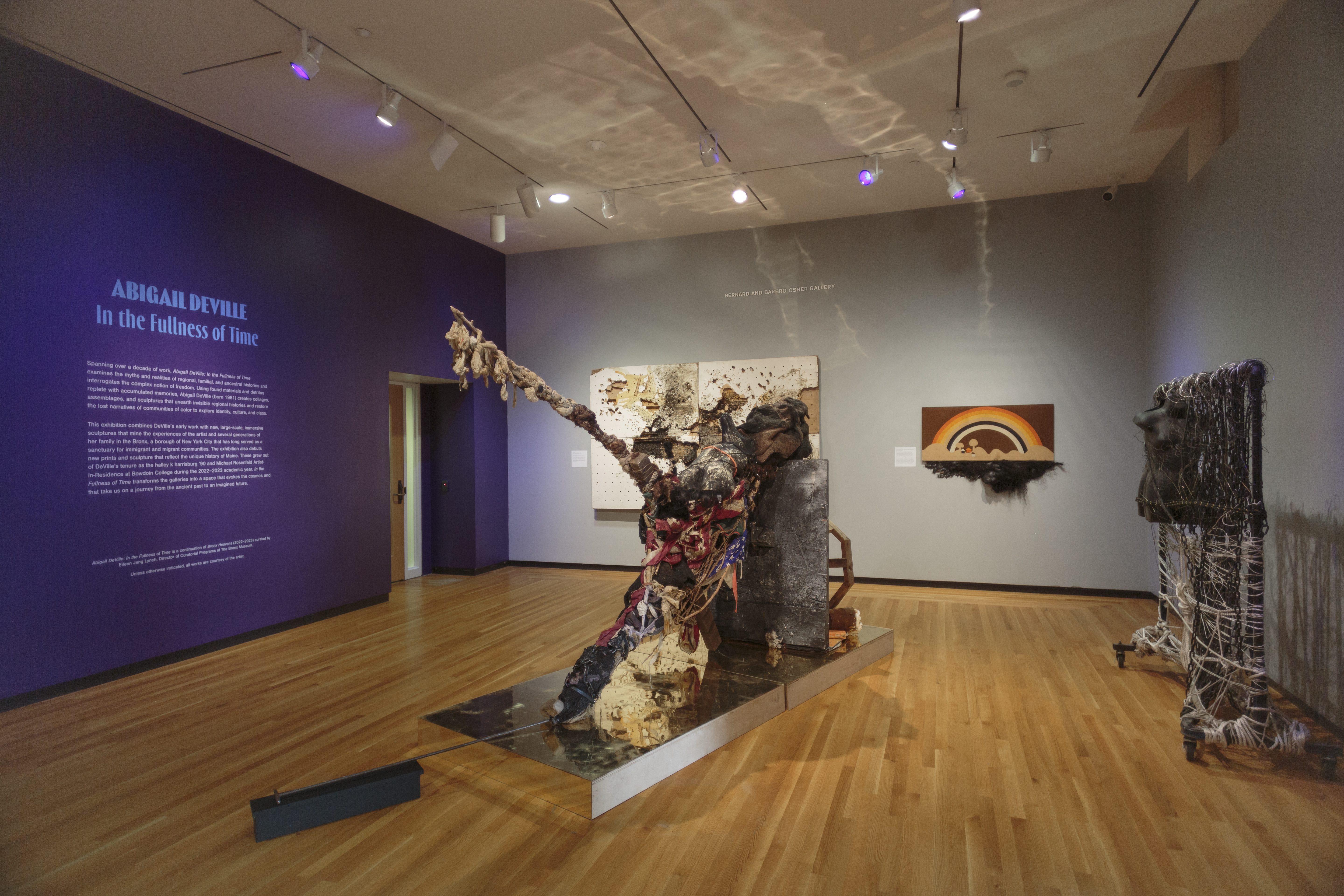 A gallery space with two gray and one purple wall displays artwork and exhibition text