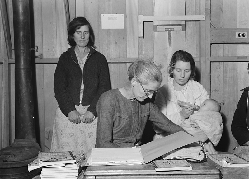 A historical black and white photograph shows six women and an infant in 1930s clothing gathered around a desk