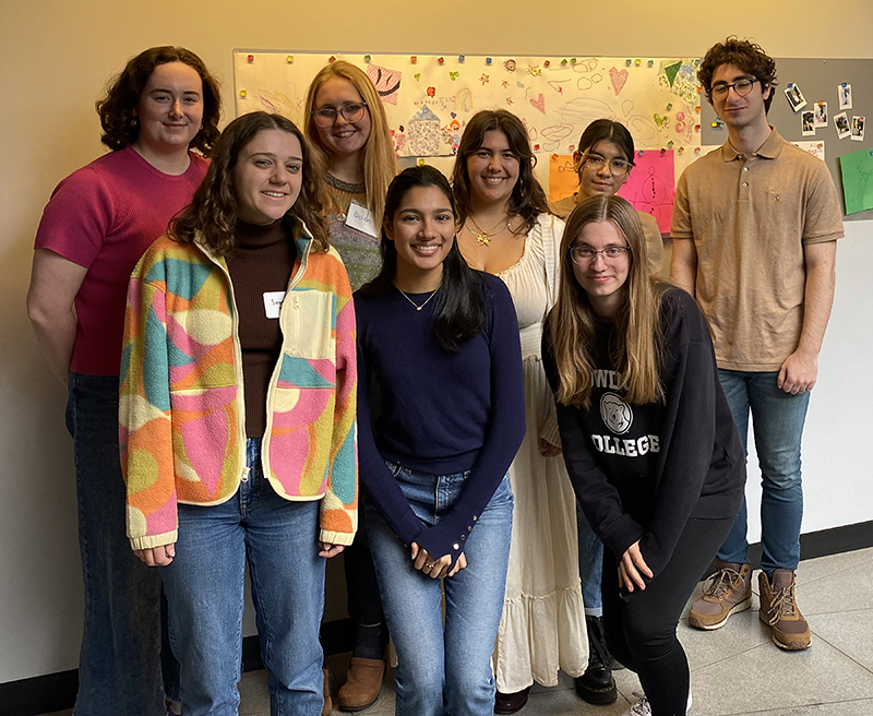 A group of 8 students posing together in a room with colorful drawings