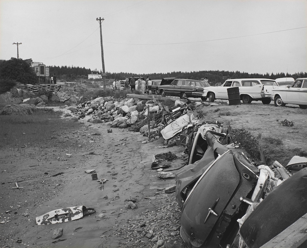 A black and white photograph shows automobile parts scattered on a beach
