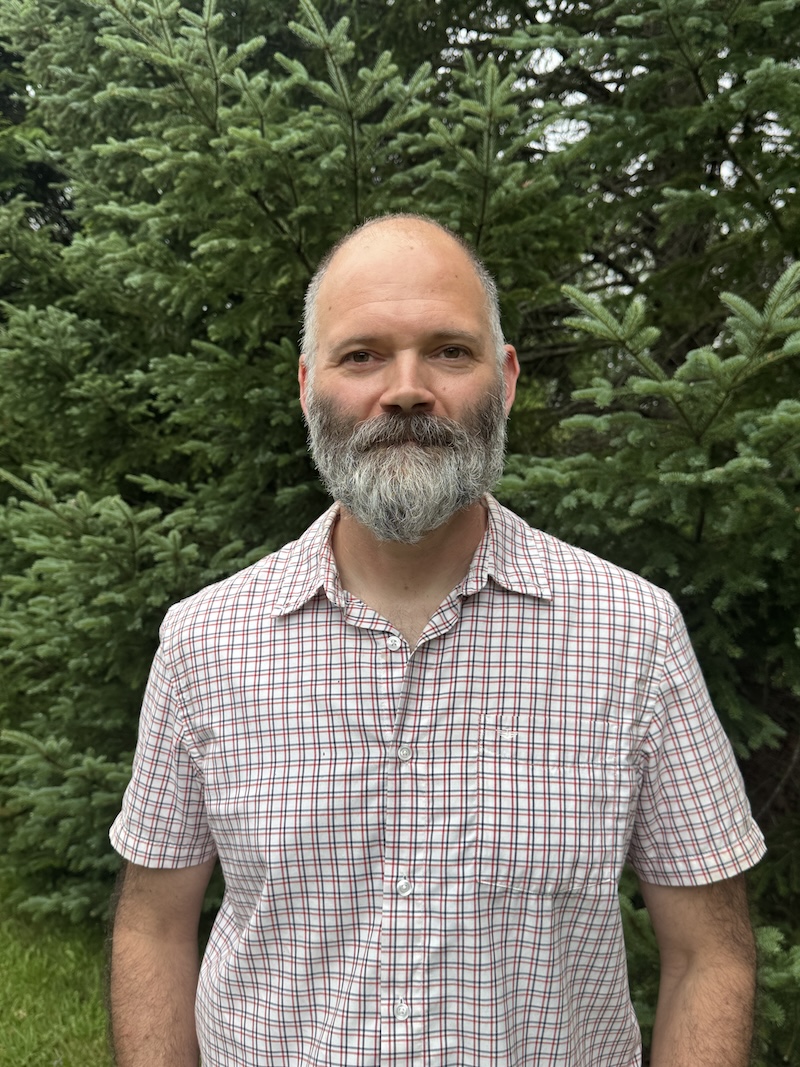 A person stands smiling with a backdrop of foliage