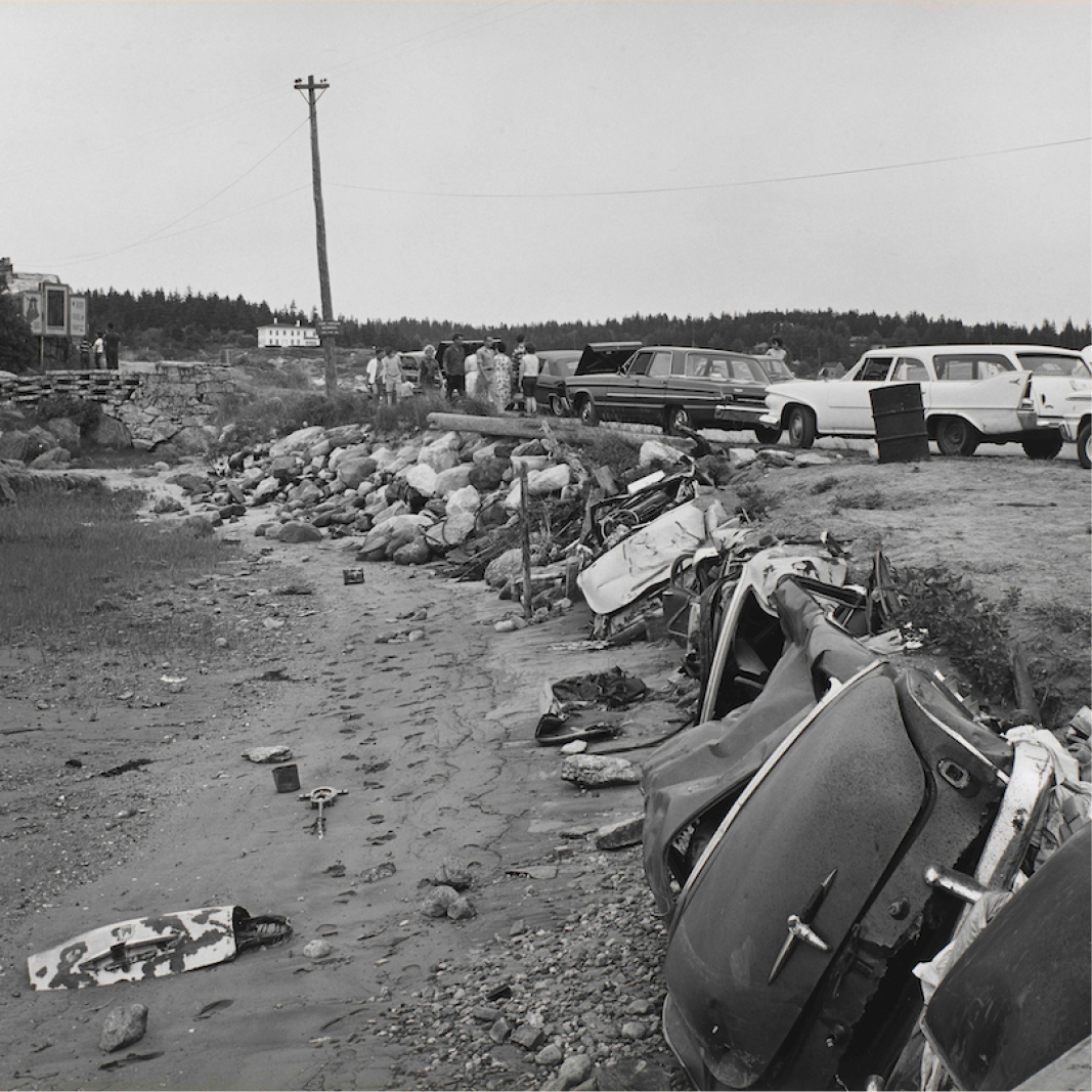 A black and white photograph shows automobile parts scattered on a beach