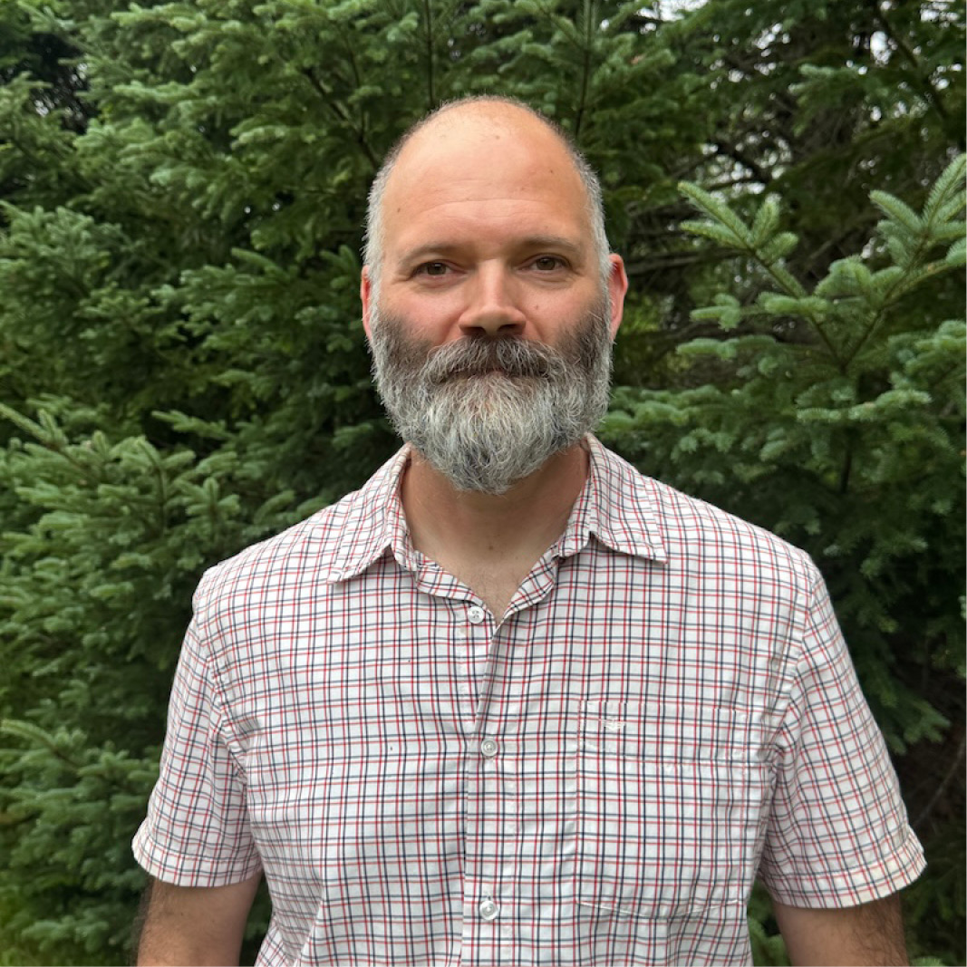 A person stands smiling with a backdrop of foliage