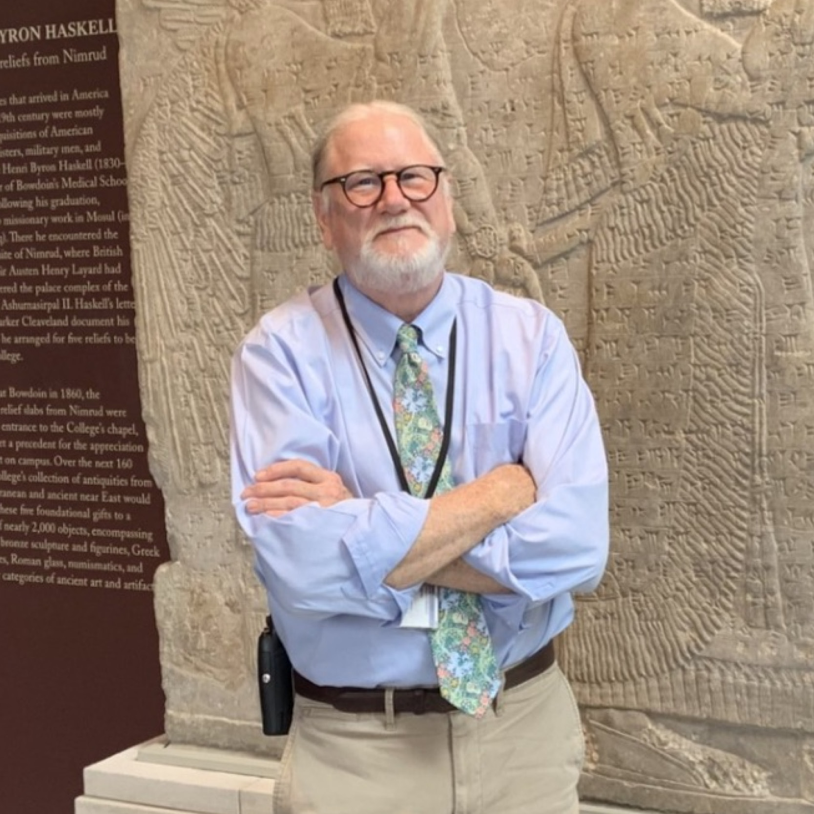 A person with glasses, a blue shirt, and tie stands in front of an Assyrian relief sculpture