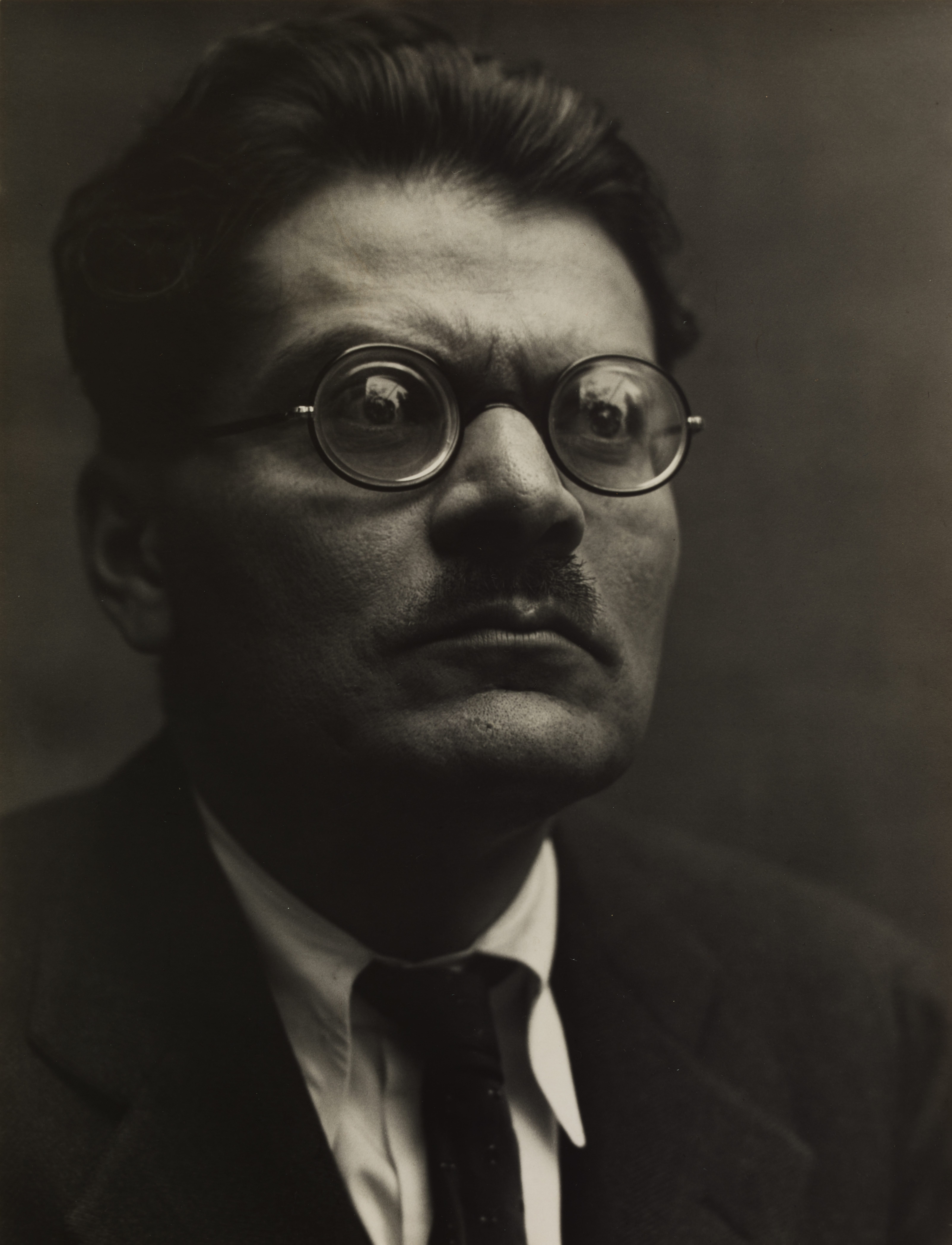 A photograph of a bust length view of a male figure wearing coke bottle glasses and a suit