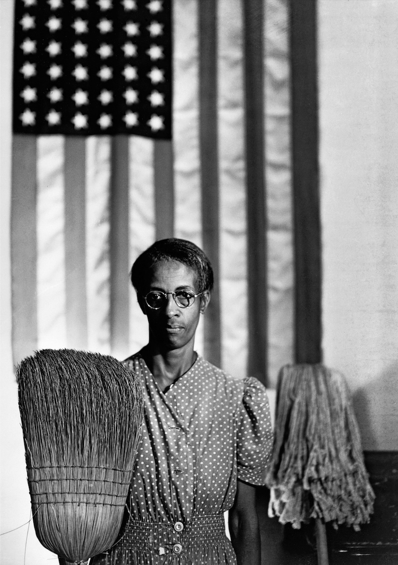 A black and white photograph of a Black woman in a polka dot shirt holding a broom and mop in front of an American flag