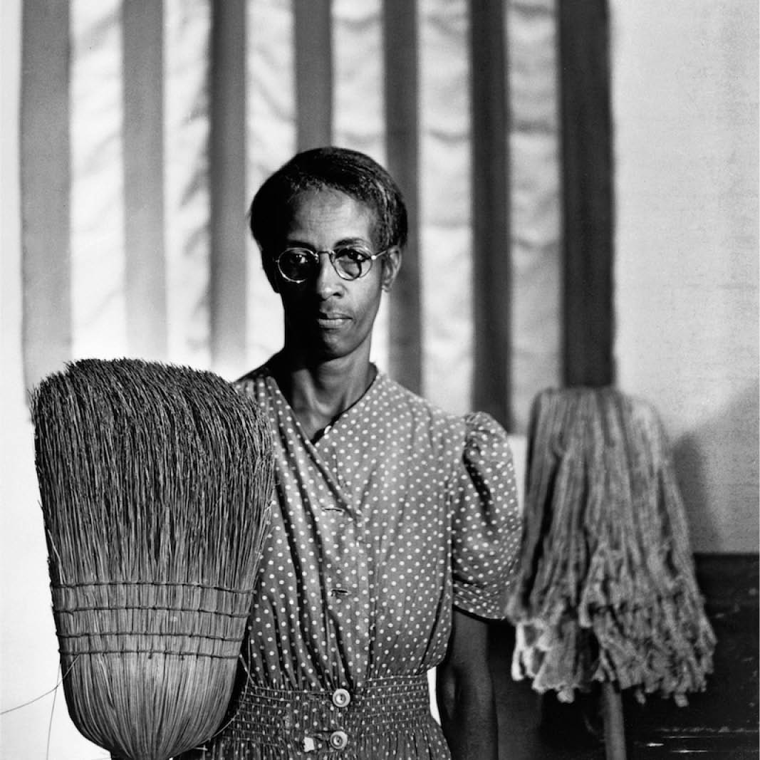 A detail of a black and white photograph of a Black woman in a polka dot shirt holding a broom and mop in front of an American flag