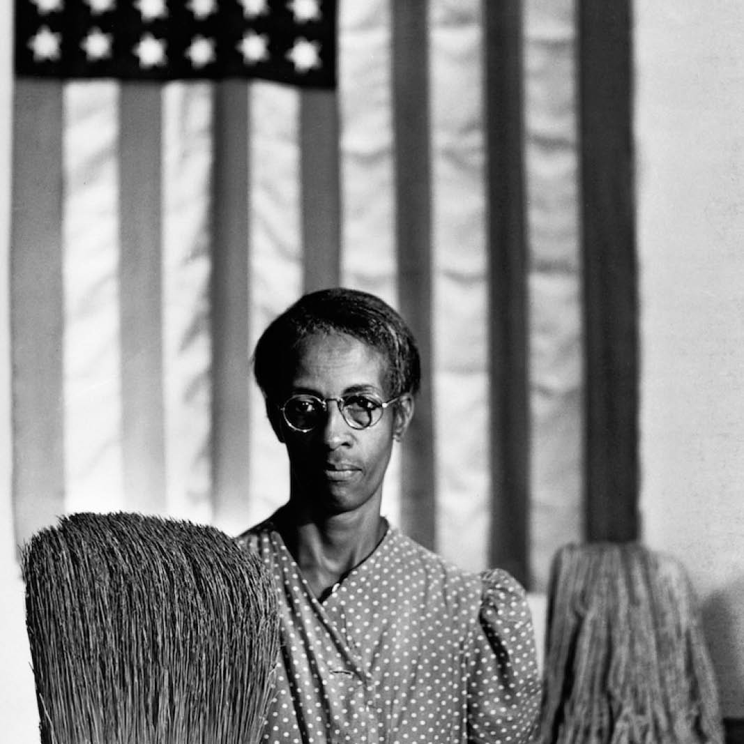 A black woman holding a broom and mop posed in front of an American flag
