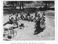 A black and white photograph of a group of people observing a cooking demonstration