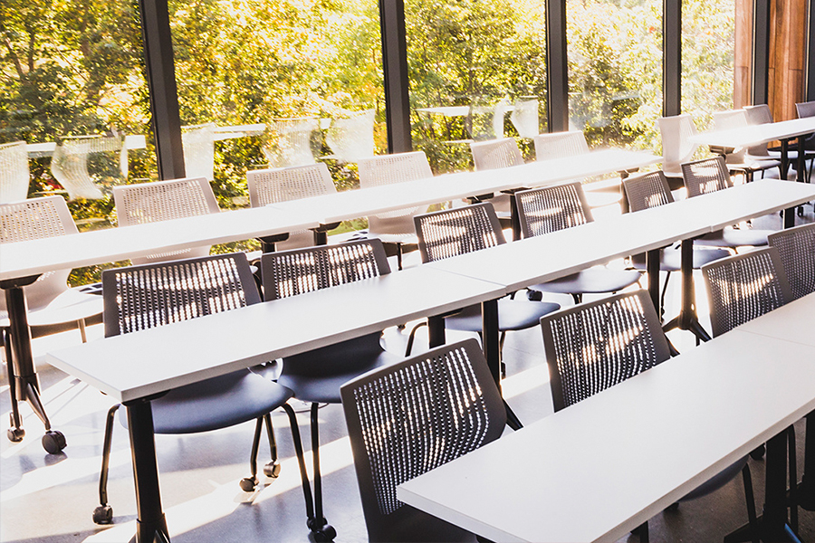 Rows of modern plasic classroom chairs behind long tables in a classroom in the Roux Center. The room has a background of large windows facing the outside.