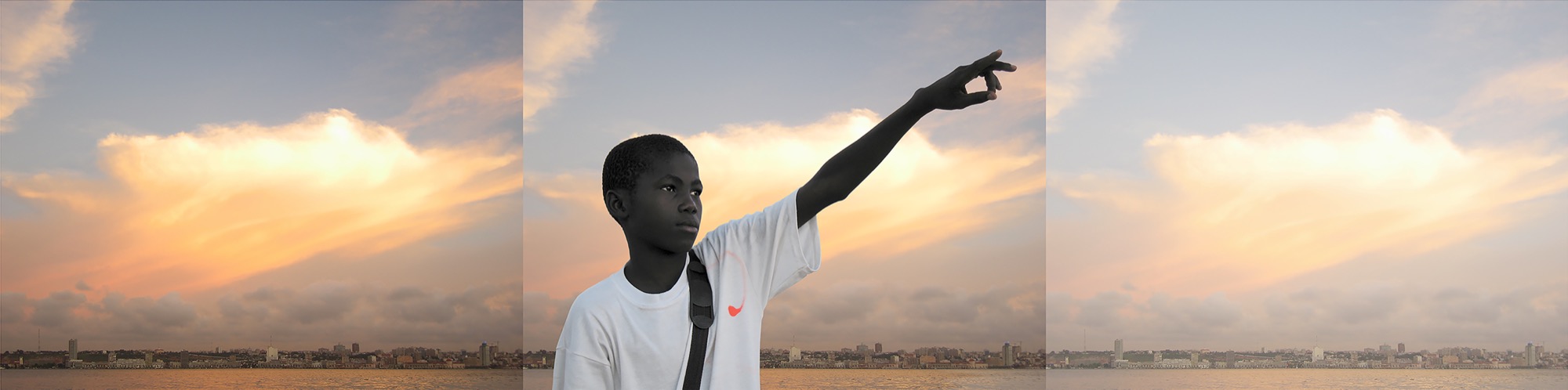a boy in a white T-shirt points upward with a city skyline behind him