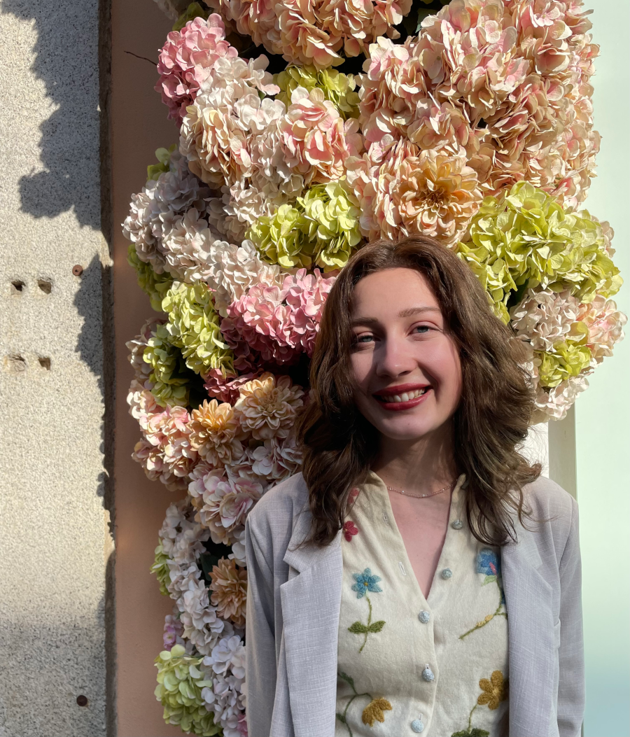 student in jacket and flower blouse stands in front of blooming flowers