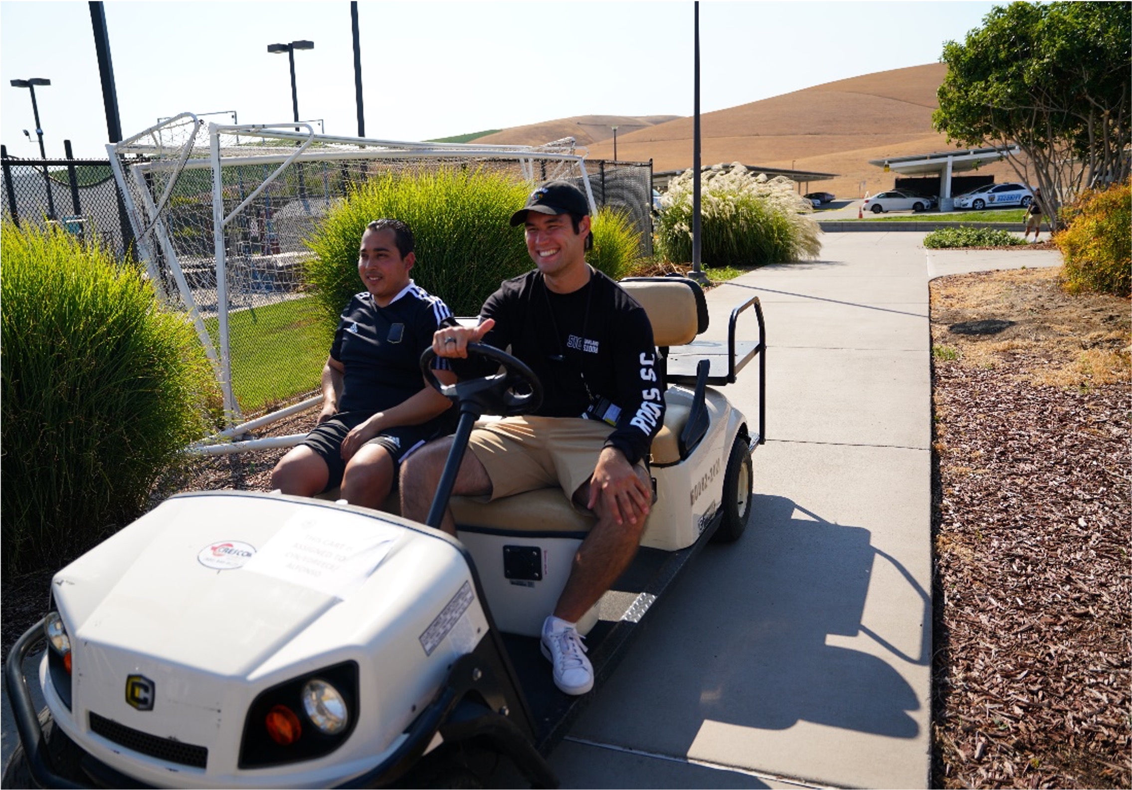 two people smiling on a golf cart in front of a soccer field