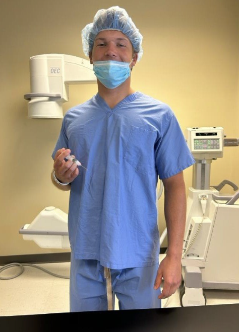 Colin poses in scrubs, holding a syringe.