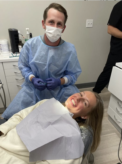 Mike is seated next to a patient, wearing dental scrubs and holding equipment.