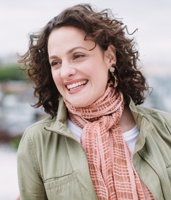 A woman with curly brown hair smiles broadly while wearing a green jacket and coral-colored patterned scarf, with a blurred waterfront background.