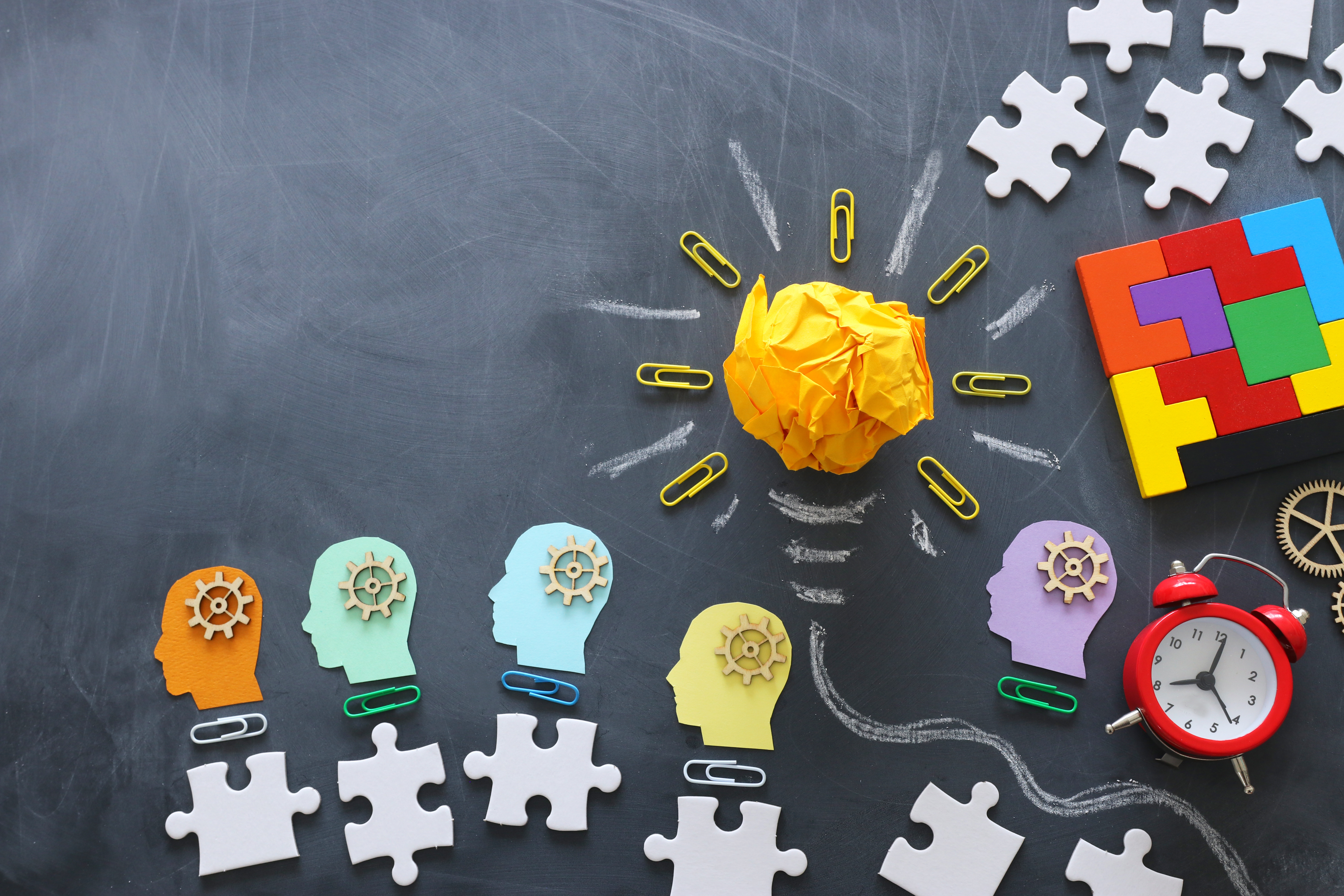 Colorful head silhouettes containing gears, puzzle pieces, and a crumpled paper light bulb on a chalkboard