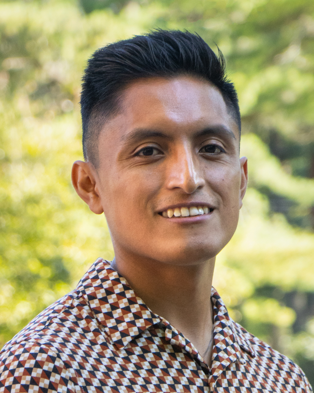 A man with short dark hair smiles while wearing a checkered shirt in black, brown, and white tones, with green foliage in the background.