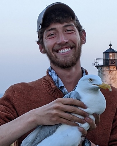 A smiling man in an orange sweater and hat holds a live seagull with a lighthouse in the background.