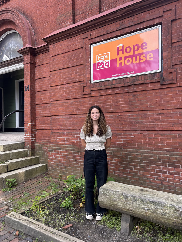 Claire stands in front of a brick building with a sign that reads "Hope House".