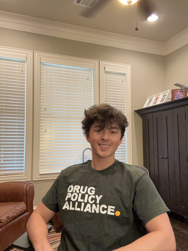 Andrew sits at a desk wearing a shirt that reads 'Drug Policy Alliance.'