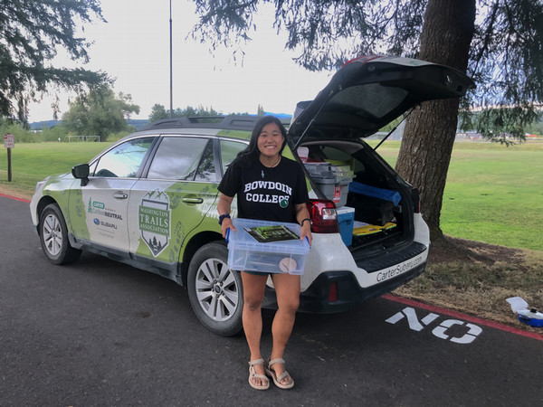 Lily stands next to a car and holds a box.