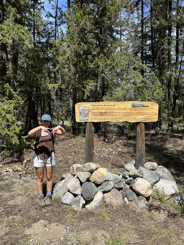 Jane stands in front of a sign in a forest, making a heart with her fingers.