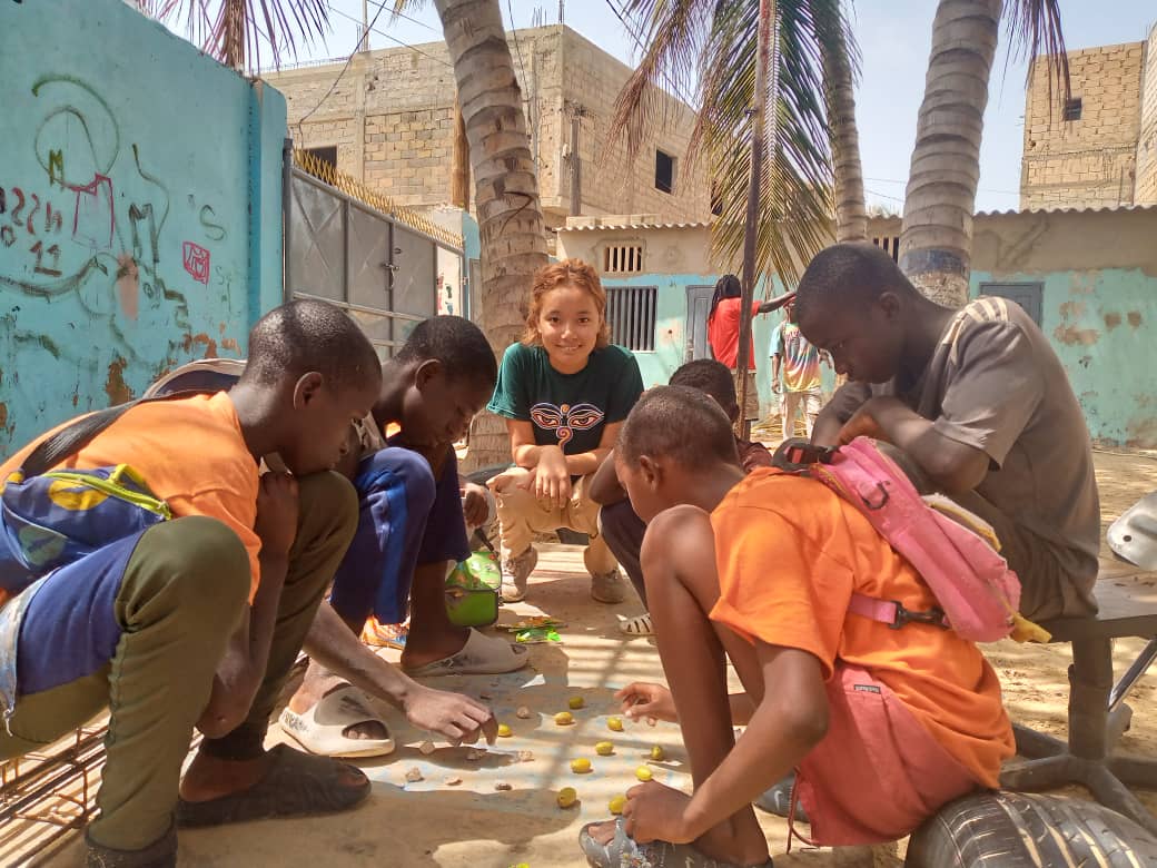 Pincho squats in an outdoor space, next to five others who are also squatting and playing a game on the floor