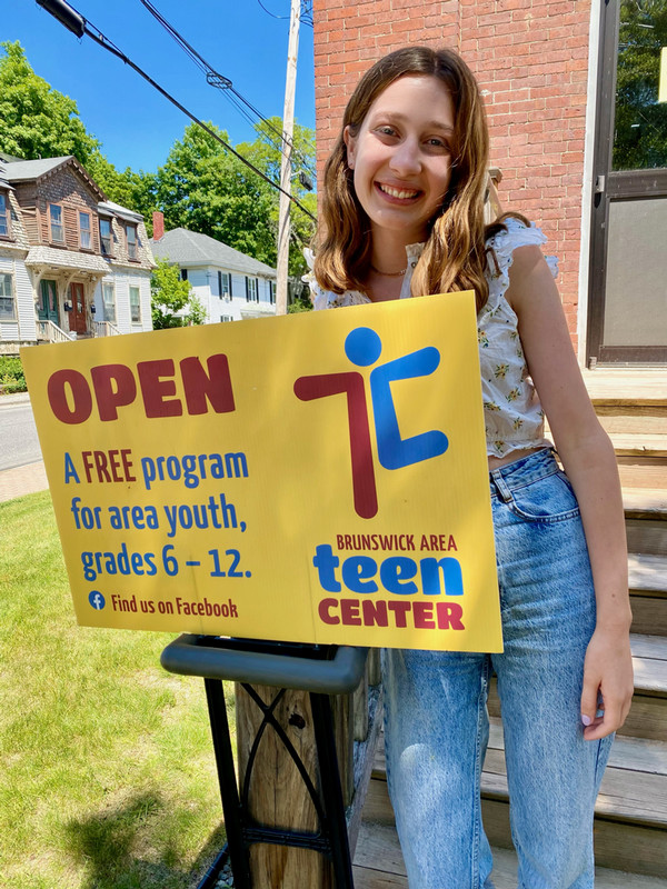 Tess stands outside of a house, with a sign in front of Tess.