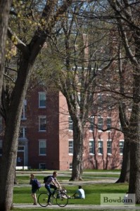 Campus in the spring; students bicycle and walk along the sidewalk; background of trees and Bowdoin building