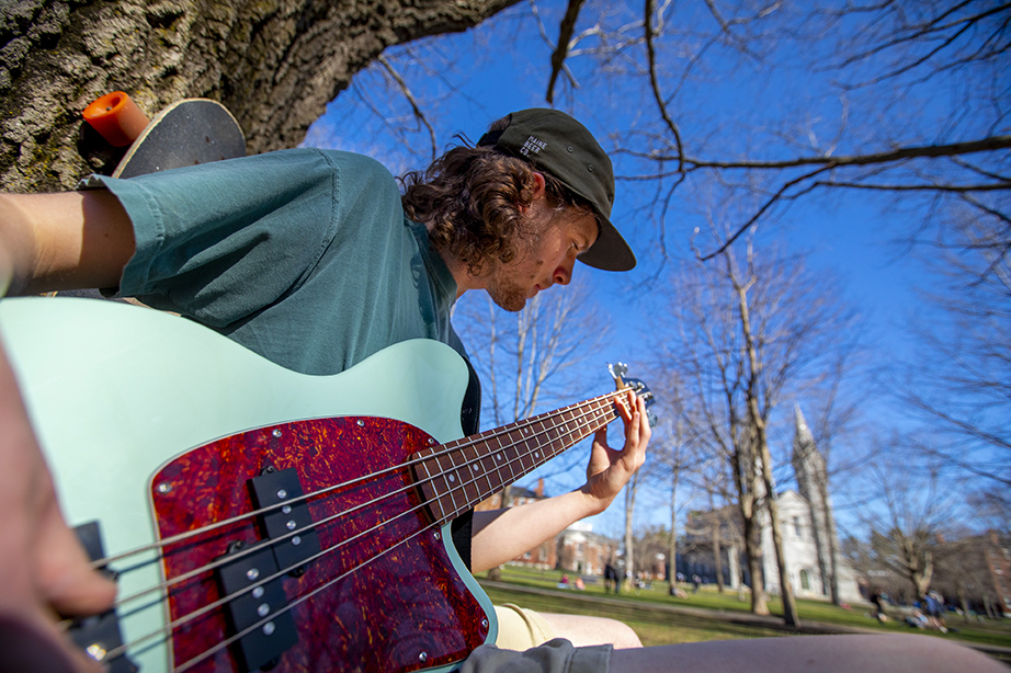 A student playing at guitar outside on the quad. The camera is very close and at a low angle, focused on the guitar and the student's hands.