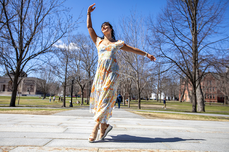 A student dancing on a walkway in the main quad. She is wearing a floral-print dress and standing on her toes, with her right arm extended above her head and her left arm extended straight out to her side at the shoulder.