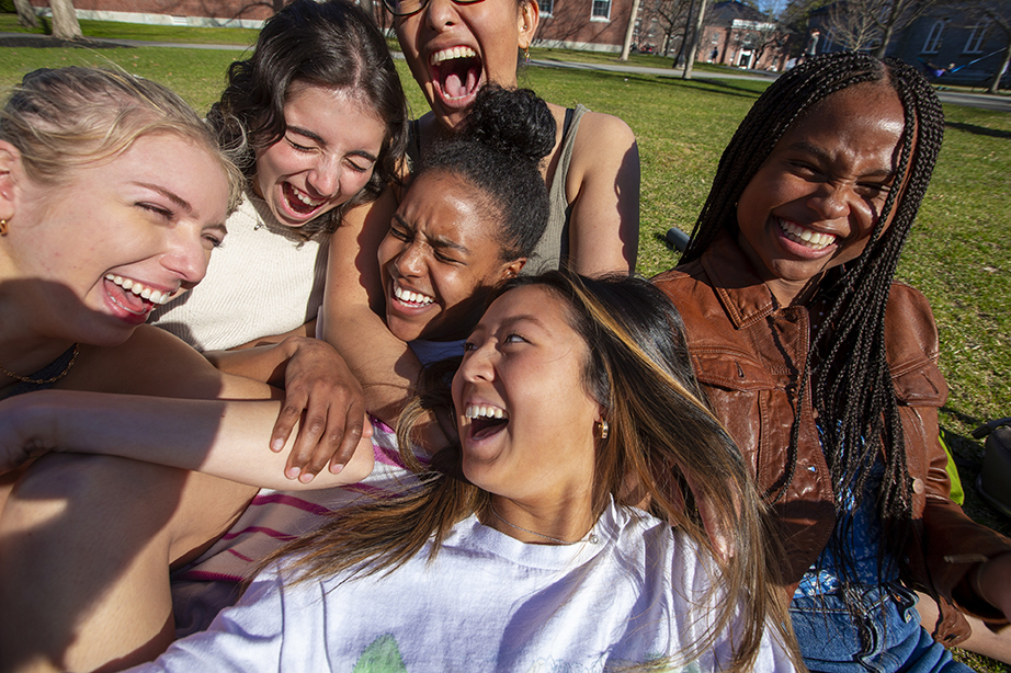 Six students sitting close to each other on the quad, laughing and holding each other.