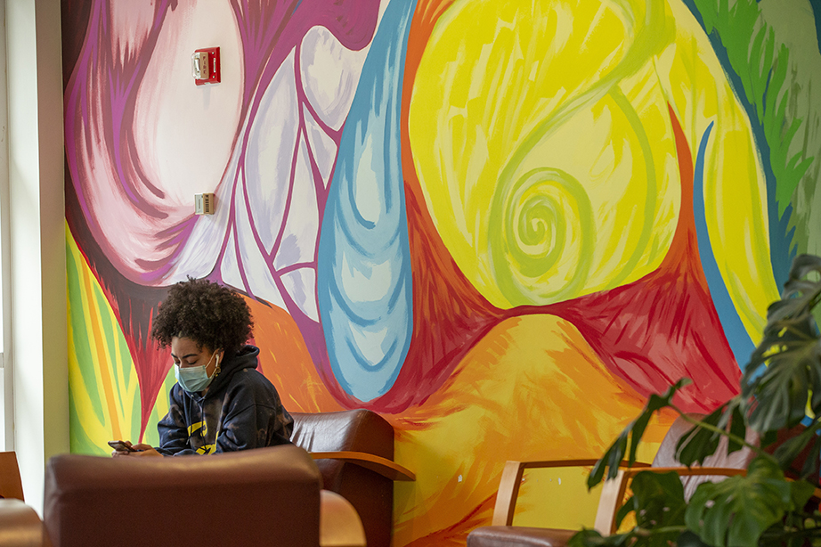 A student sitting on a couch in front of an intricate, brightly colored mural of swirling curves and natural shapes.