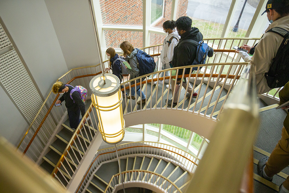 Several students descending a spiral staircase in a building with light streaming in from a nearby window.