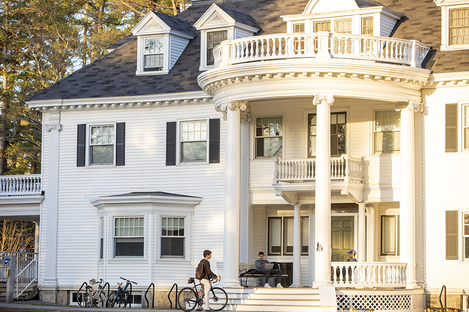 Two students sitting on the round porch of a large, white building. They are talking to a third student, who is standing nearby with his bike.