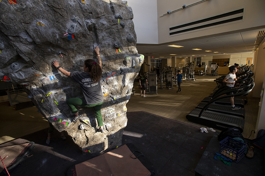 A student climbing the rock wall in the Buck Center, partially illuminated by light streaming in through the window.