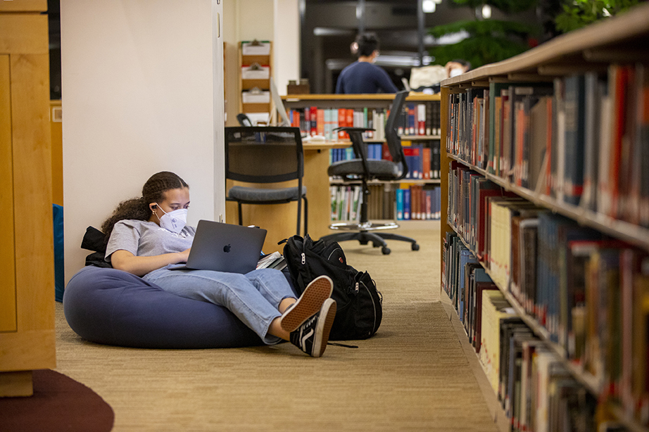 A student sitting on a bean bag across from a library bookshelf, looking at a laptop on her lap.