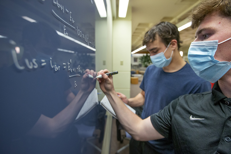 A close shot of two students standing in front of a black markerboard. The student in the foreground is about to write down something with a white marker, while the student in the background looks at a piece of paper in his hands.