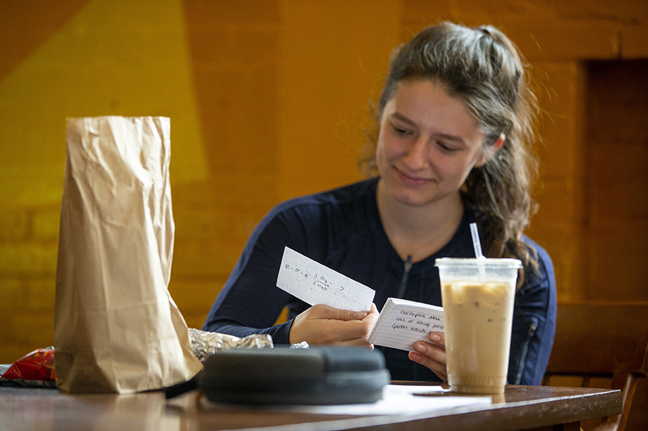 A student sitting at a table studying handwritten flashcards, a brown paper bag and iced coffee in front of her.