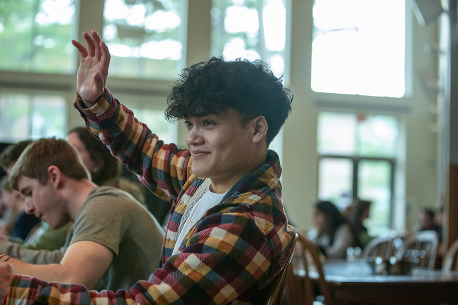 A student sitting at a table in Thorne dining hall raising a hand in greeting to someone out of frame.