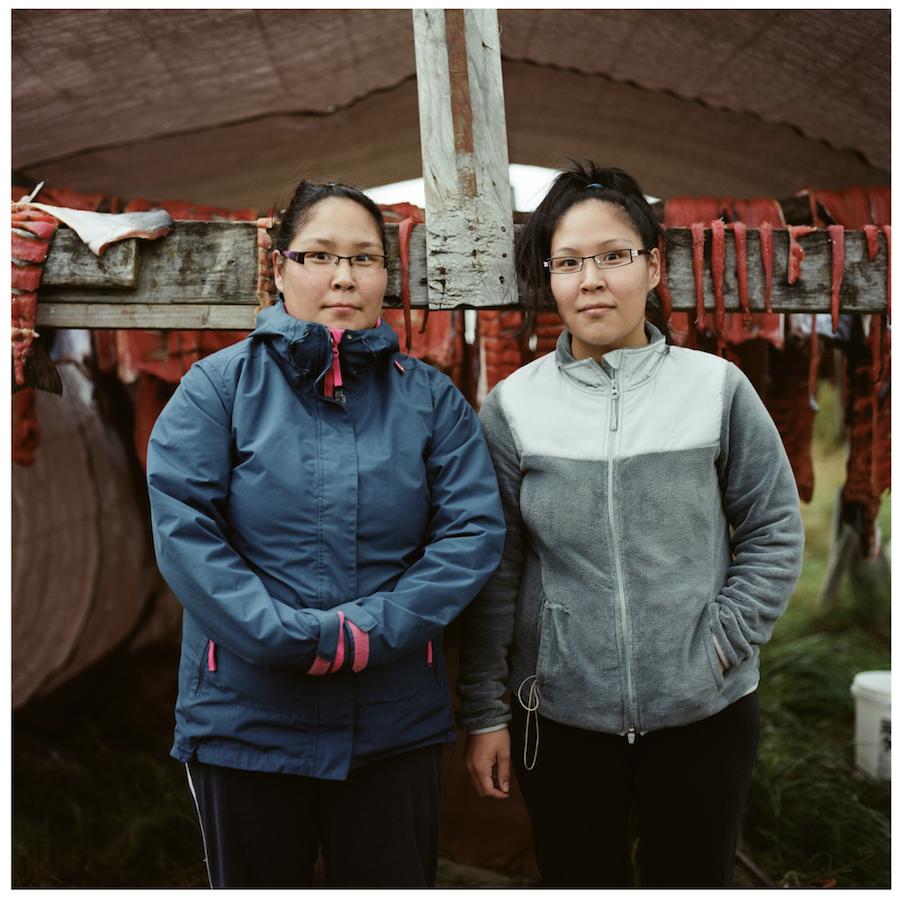 Britany Cleveland and Taryn Andrew are standing in front of a traditional Yup'ik fish rack full of silver salmon drying after a successful fishing season, Brian Adams, 2015. Courtesy of the artist.